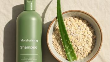 Flatlay av fuktgivende shampoo med keramisk skål havregryn og fersk aloe vera-blad på krem lin, varmt dagslys